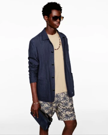 A young Black man poses in a casual outfit, featuring a beige T-shirt, patterned shorts, a navy jacket, sunglasses, and a beaded necklace.