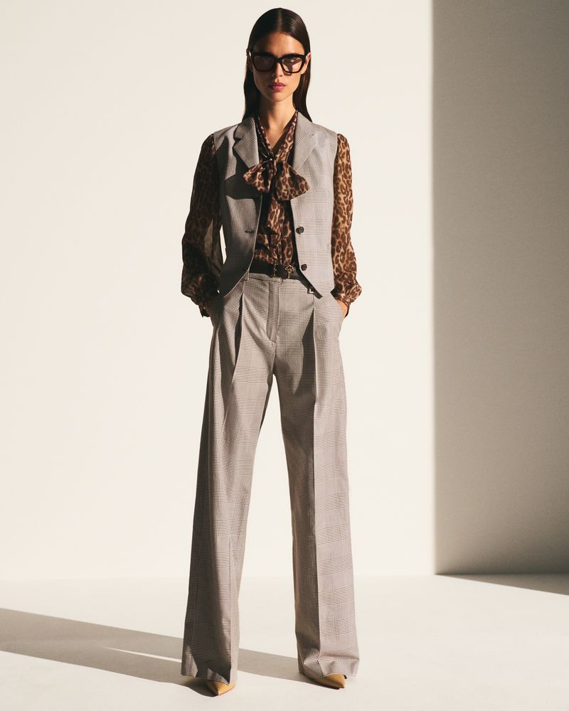 A woman wearing a grey vest, brown patterned blouse, grey trousers and black framed glasses poses in a bright photo studio.