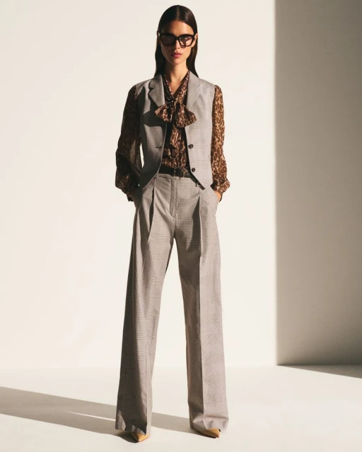 A woman wearing a grey vest, brown patterned blouse, grey trousers and black framed glasses poses in a bright photo studio.