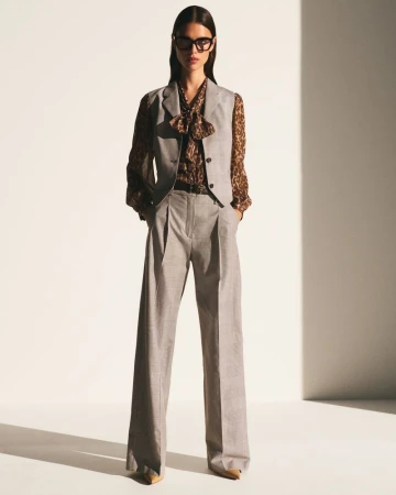 A woman wearing a grey vest, brown patterned blouse, grey trousers and black framed glasses poses in a bright photo studio.