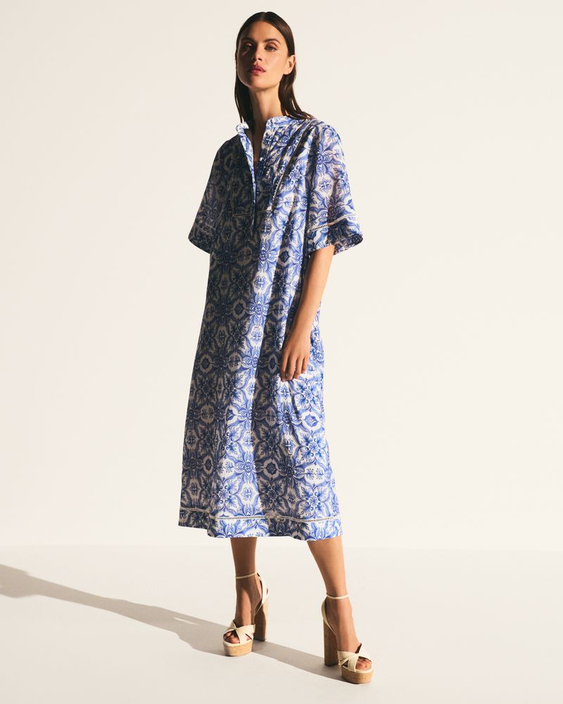 A slender brunette woman poses in a full-length studio shot, wearing a blue and white printed midi dress and platform heels.