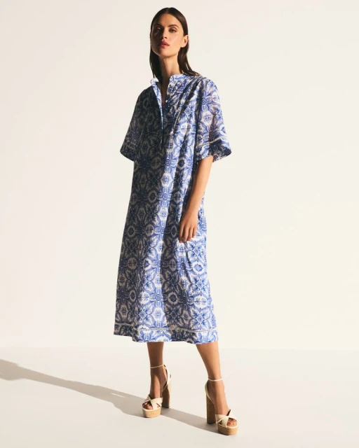 A slender brunette woman poses in a full-length studio shot, wearing a blue and white printed midi dress and platform heels.