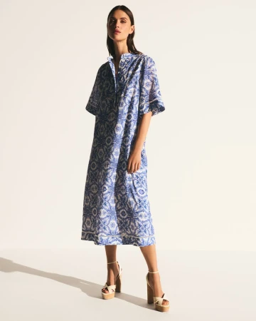 A slender brunette woman poses in a full-length studio shot, wearing a blue and white printed midi dress and platform heels.