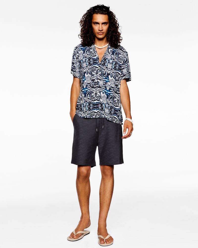 Male fashion model stands against a white backdrop, wearing a patterned short-sleeve shirt, dark shorts, and neutral flip-flops.