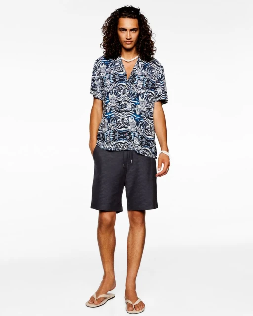 Male fashion model stands against a white backdrop, wearing a patterned short-sleeve shirt, dark shorts, and neutral flip-flops.