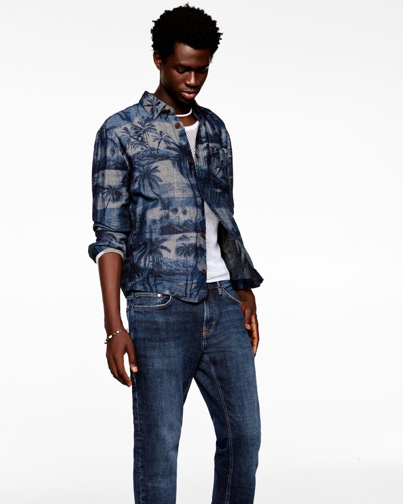 An adult male is standing in a photo studio, wearing a long-sleeved palm-tree-patterned shirt, a white undershirt, and blue jeans.