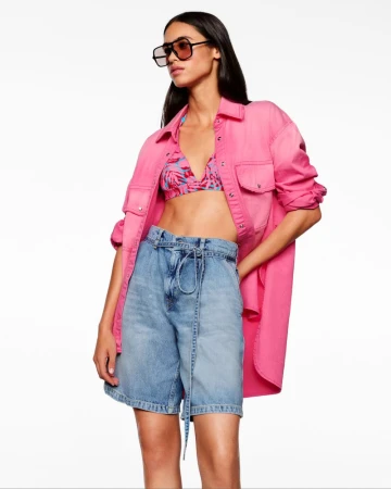 Brunette woman in pink button-up shirt, bikini top, denim shorts, and sunglasses posing in front of a white background.