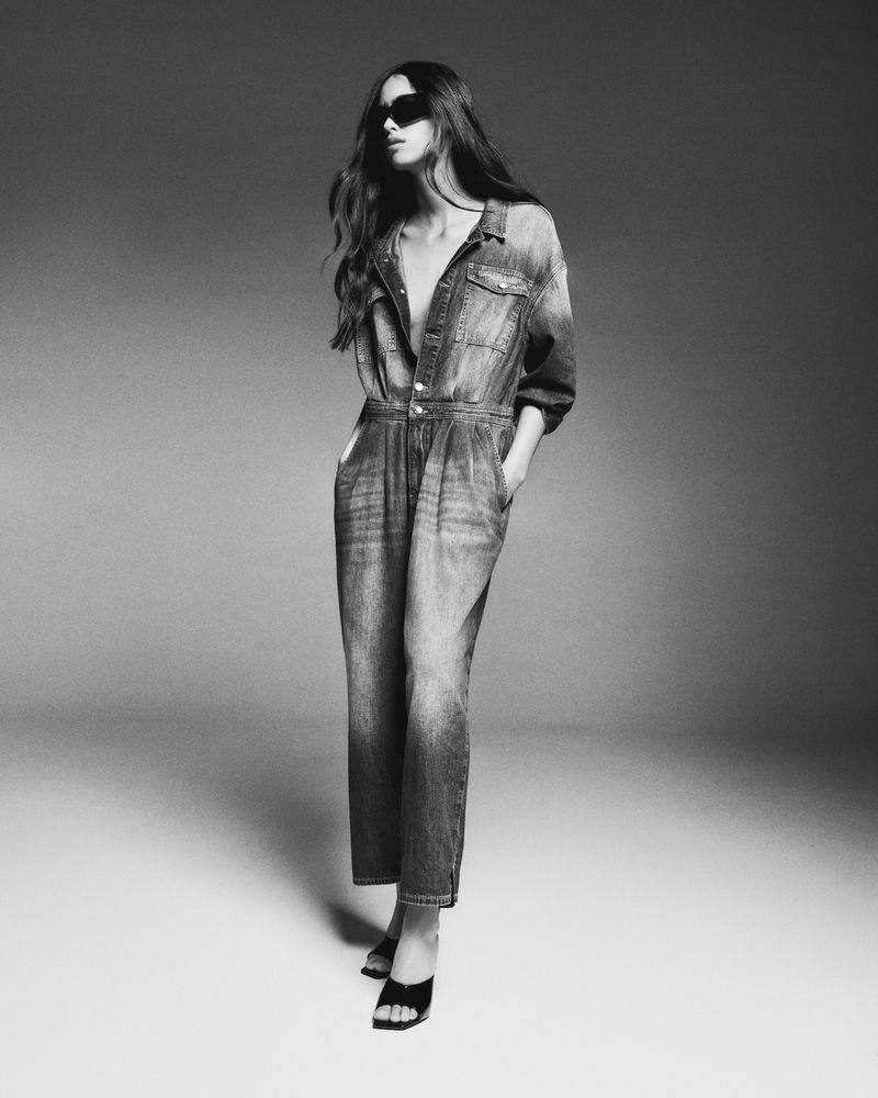 A woman in a denim jumpsuit and black sunglasses poses in a studio with neutral background, standing with hands in her pockets.