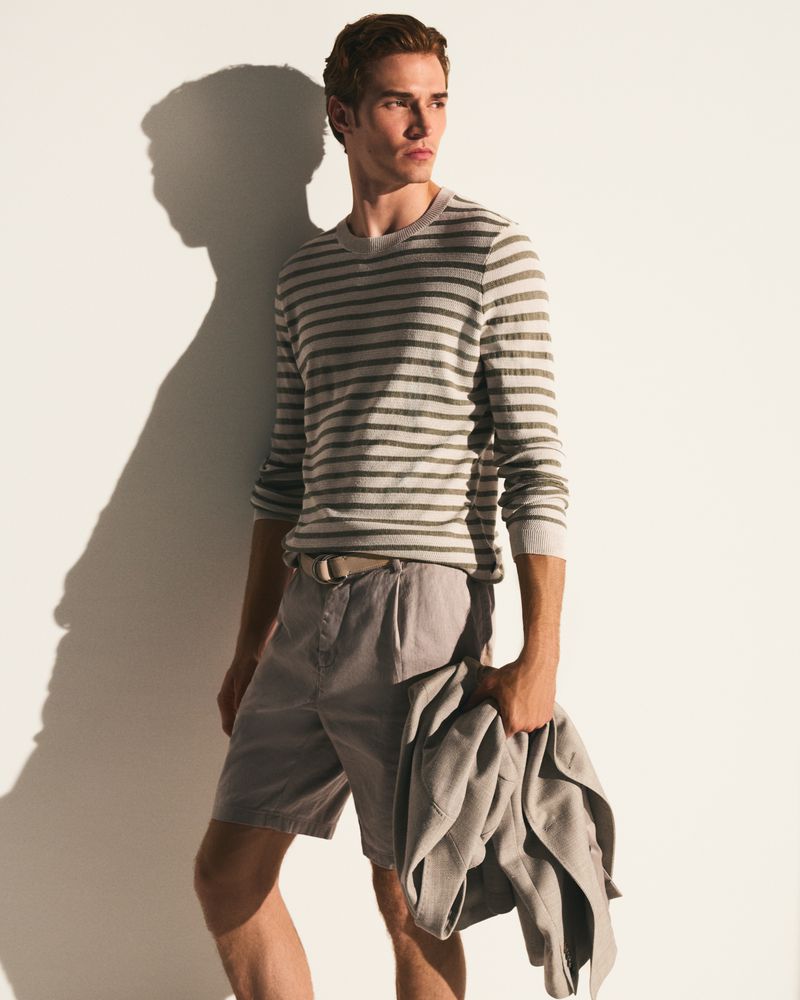 Man in striped shirt and shorts holding a jacket, standing against a white wall with a shadow. Fashion editorial photography.