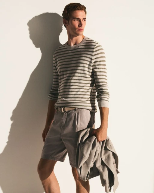 Man in striped shirt and shorts holding a jacket, standing against a white wall with a shadow. Fashion editorial photography.
