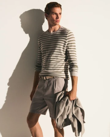 Man in striped shirt and shorts holding a jacket, standing against a white wall with a shadow. Fashion editorial photography.