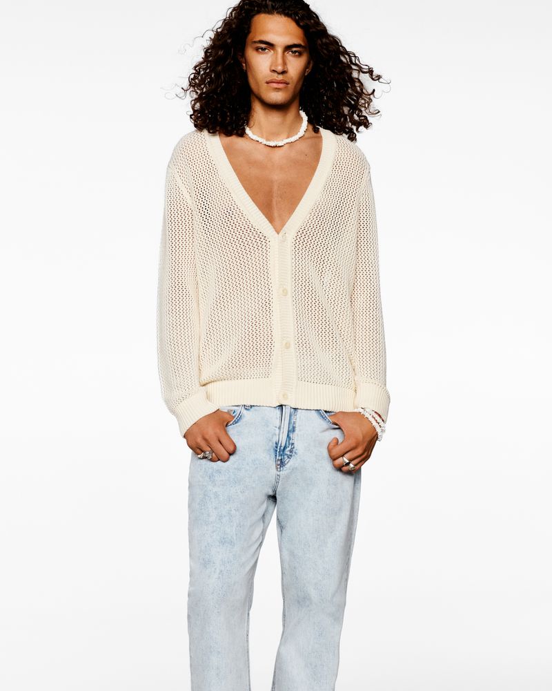 Fashion portrait of an adult male model wearing a white knit cardigan, jeans, and a pearl necklace against a plain white background.