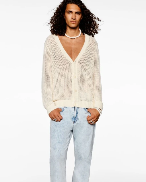 Fashion portrait of an adult male model wearing a white knit cardigan, jeans, and a pearl necklace against a plain white background.