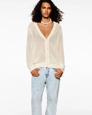 Fashion portrait of an adult male model wearing a white knit cardigan, jeans, and a pearl necklace against a plain white background.