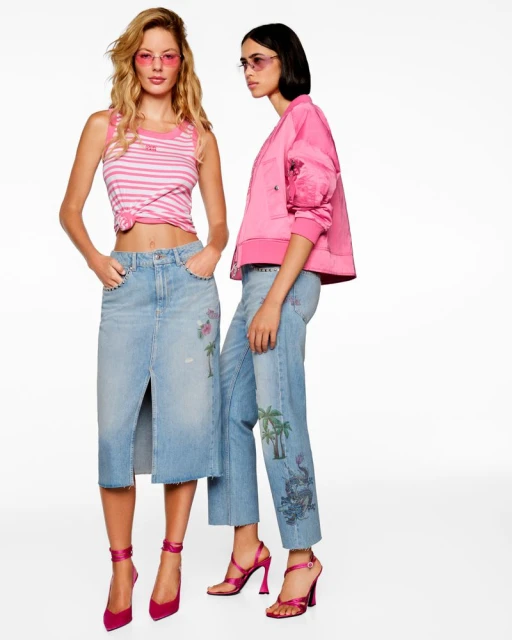Two women in pink sunglasses pose wearing denim skirts and jeans with pink tops and jackets in a brightly lit photo studio.
