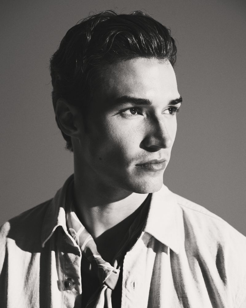 Monochrome portrait of a handsome adult male with short, wavy dark hair, wearing a striped shirt and jacket, looking away from the camera.