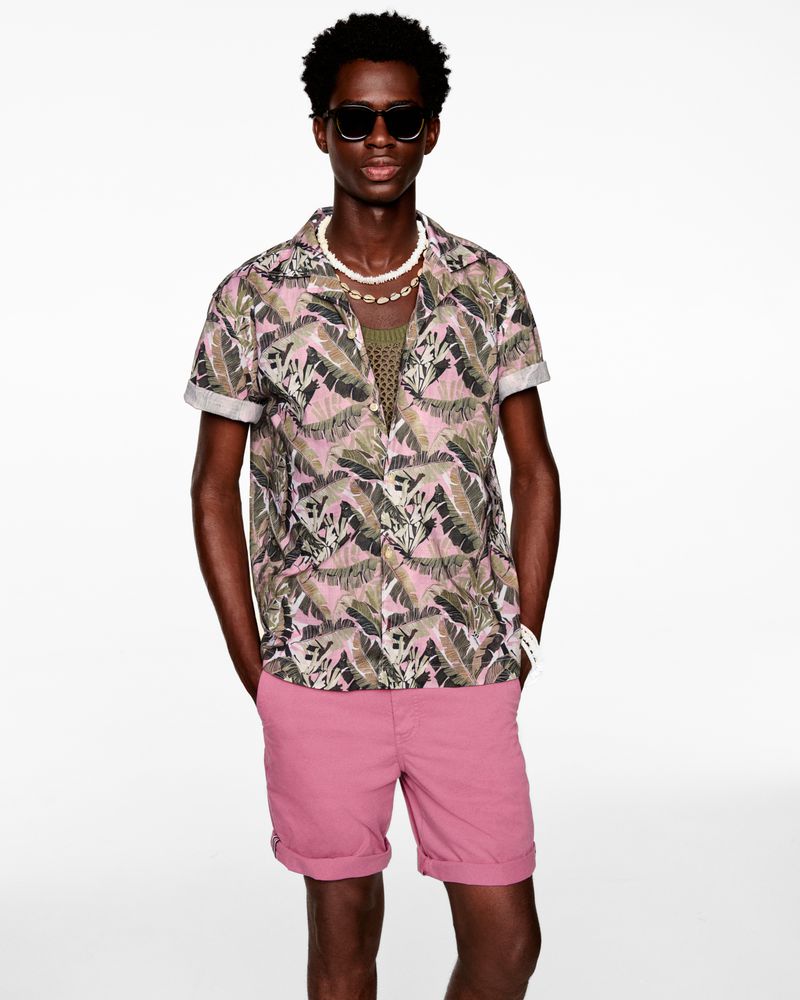 A dark-skinned man in a tropical print shirt and pink shorts poses against a white background, wearing sunglasses and a shell necklace.