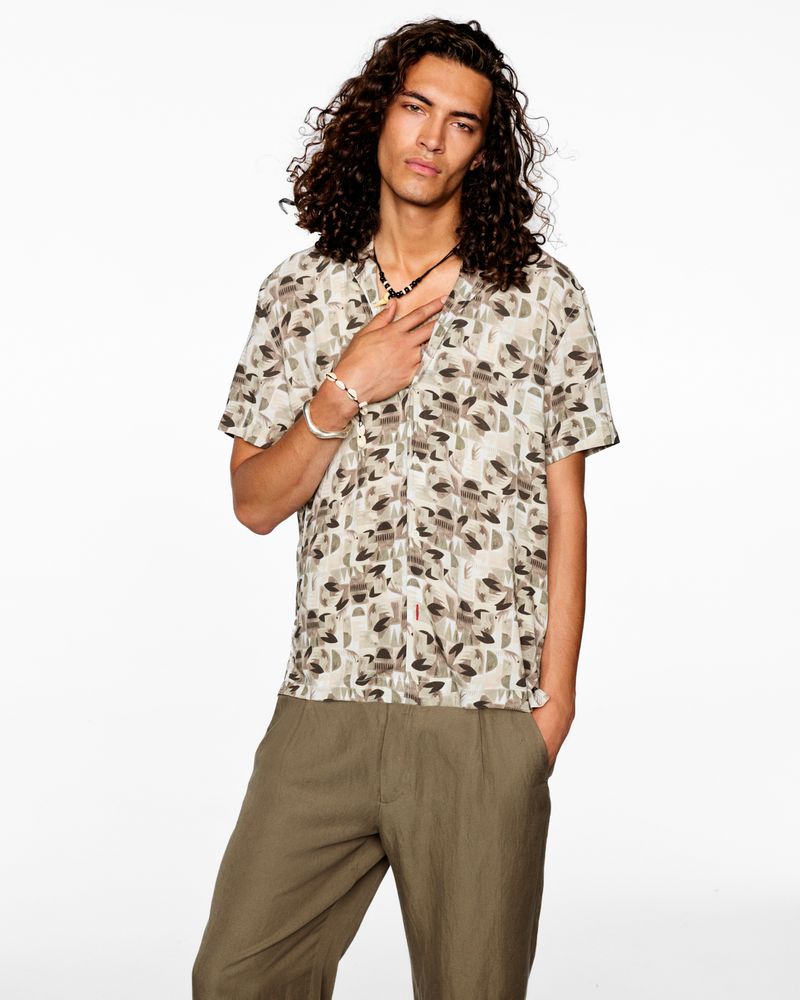 Portrait of a young adult male with curly hair wearing a patterned shirt and pants against a white backdrop in a studio setting.