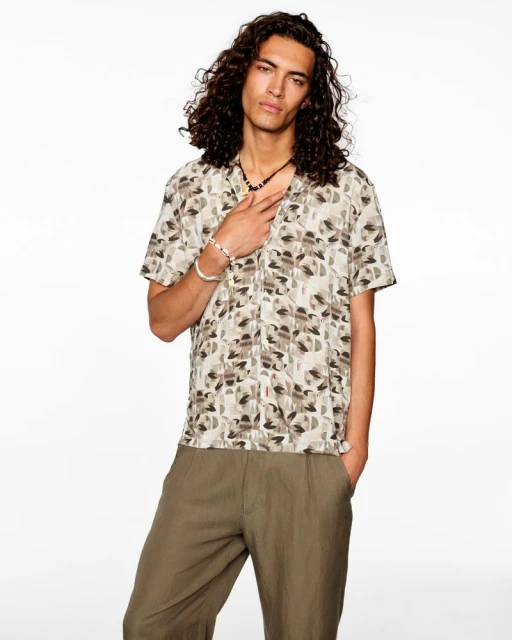 Portrait of a young adult male with curly hair wearing a patterned shirt and pants against a white backdrop in a studio setting.
