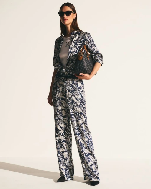 Woman in patterned pantsuit holding satchel handbag, standing in photo studio wearing sunglasses. Fashionable look with neutral background.