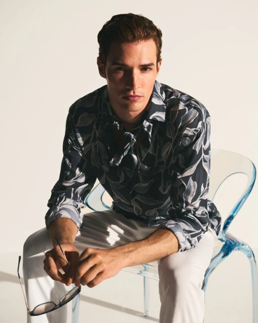 Portrait of an adult male in a patterned shirt and white pants sitting on a blue transparent chair in a bright photo studio.