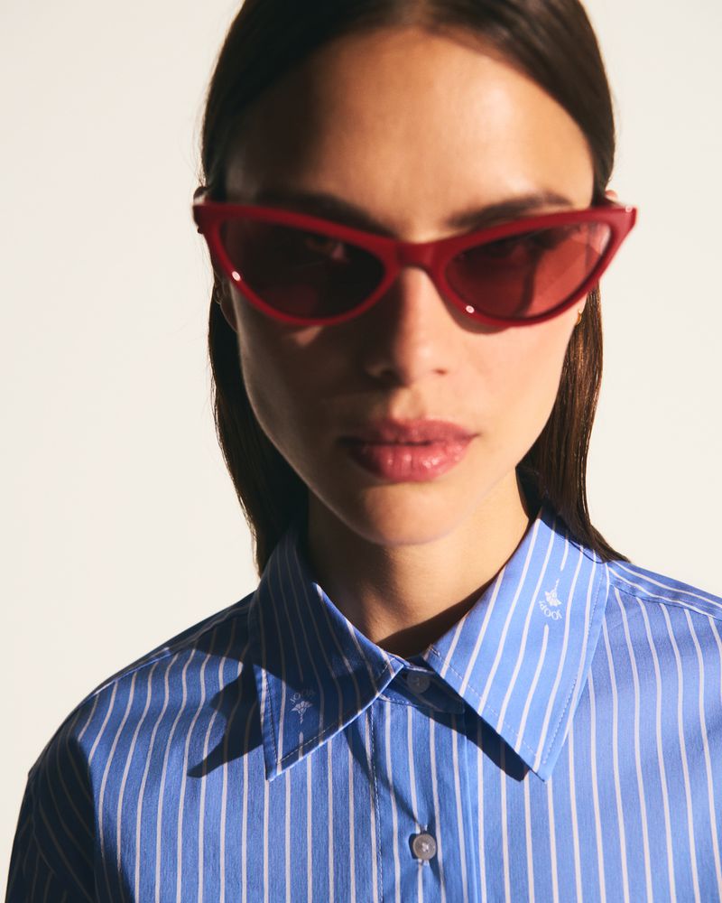 Close-up of a woman wearing a striped blue shirt and red cat-eye sunglasses against a white background in a studio setting.
