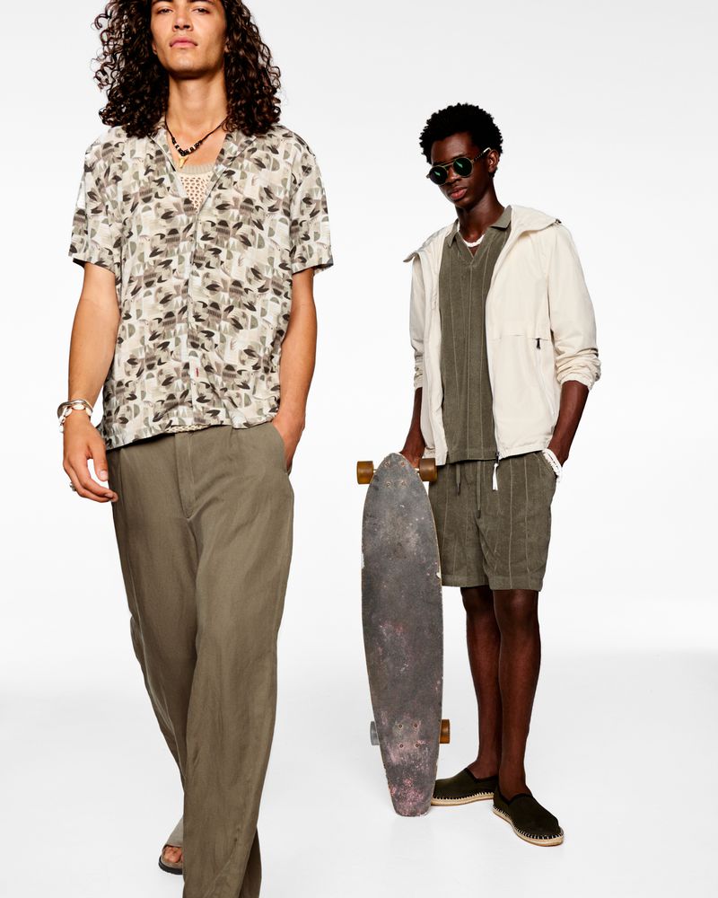 Two adult men pose in neutral casualwear against a white backdrop, one with a skateboard, showcasing modern fashion and style in a studio setting.
