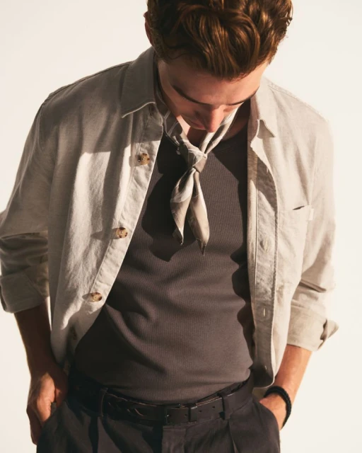 An adult male with red hair wears a gray shirt, tan jacket, dark pants and a scarf tied around his neck in a studio environment.