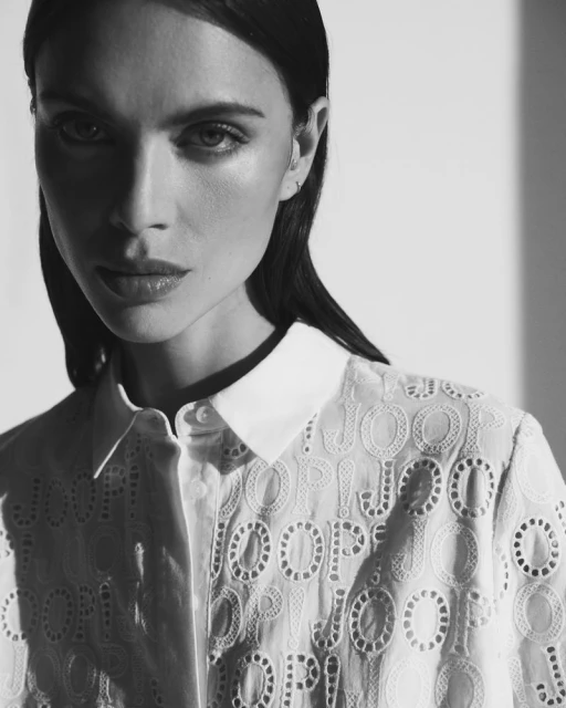 Monochrome portrait of a stylish woman in a patterned collared shirt, posing in a photo studio with light and shadow.