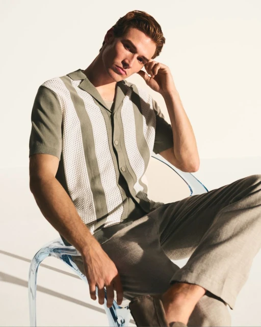 Adult male model poses in a vertical studio shot, wearing a patterned shirt and neutral-toned pants while sitting on a clear acrylic chair.