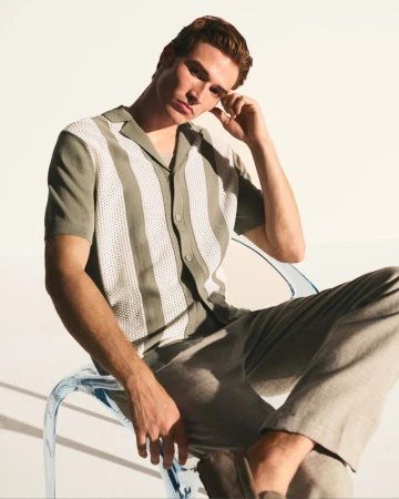 Adult male model poses in a vertical studio shot, wearing a patterned shirt and neutral-toned pants while sitting on a clear acrylic chair.