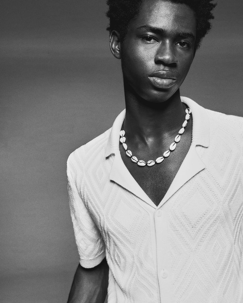 Black and white close-up of a Black male model in a diamond-patterned white polo shirt and shell necklace, against a solid gray backdrop.