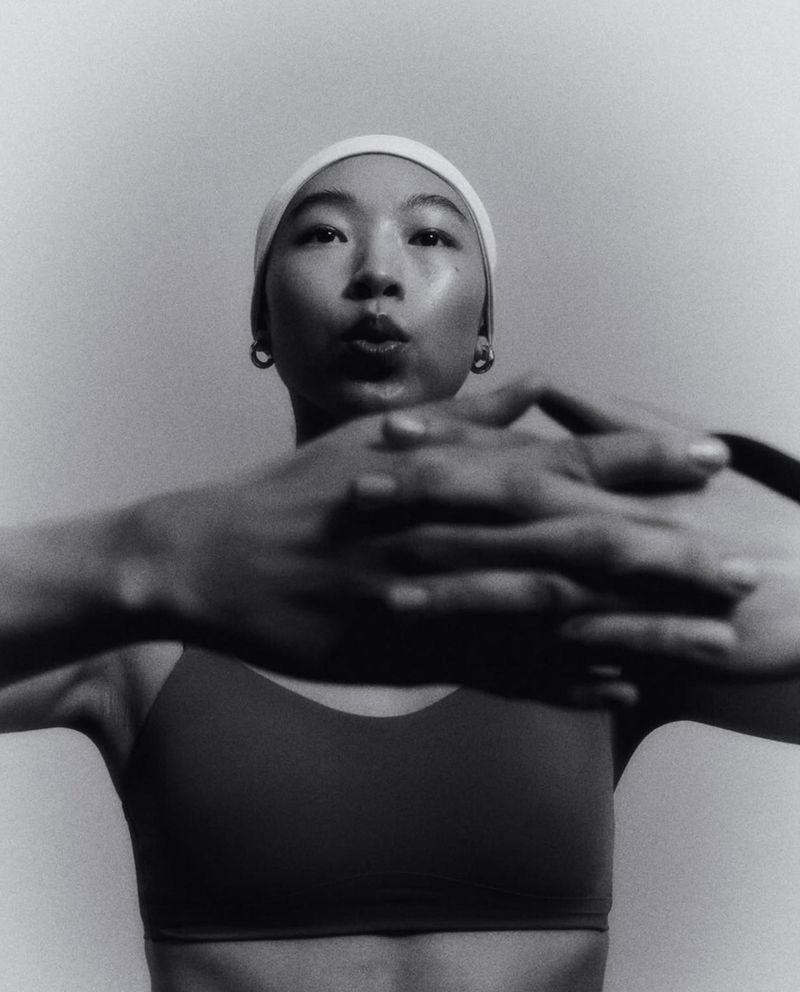 Monochromatic portrait of an adult woman with a neutral expression wearing a swim cap, earrings, and sports bra in a studio setting.