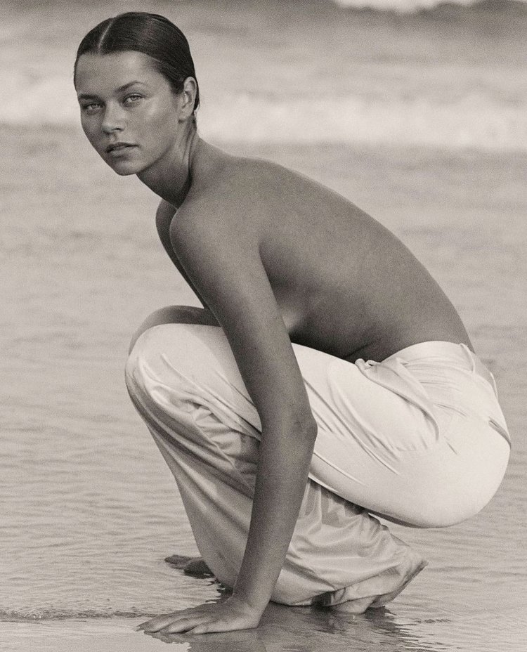 Model crouching on sandy beach wearing light-colored trousers with ocean waves in background.