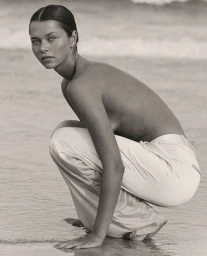 Model crouching on sandy beach wearing light-colored trousers with ocean waves in background.