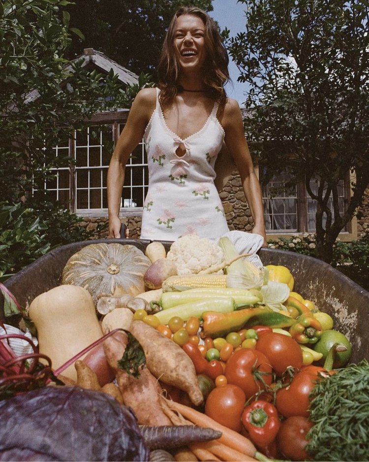 Smiling woman stands behind wheelbarrow full of fresh vegetables in a rural garden setting with a stone cottage in the background.