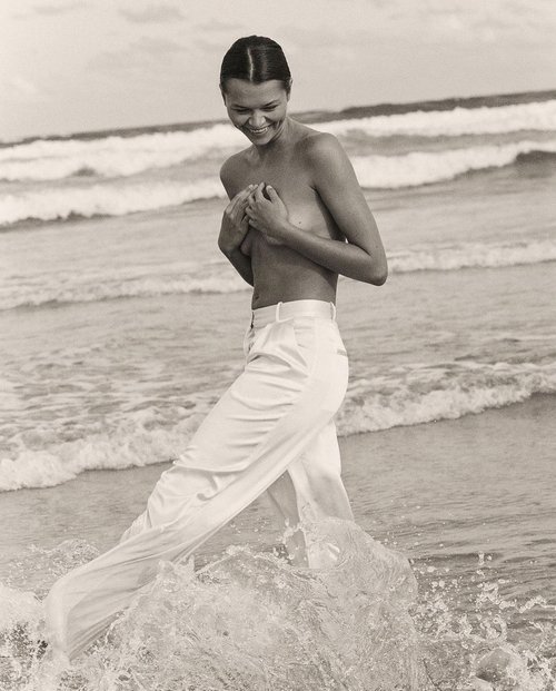 Black-and-white shot of a smiling adult woman in a landscape setting, wearing white pants while wading in the ocean with breaking waves.
