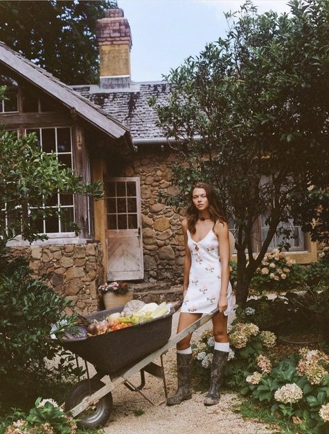 Woman in a dress pushes a wheelbarrow filled with vegetables in a garden next to a stone cottage on a sunny day, rural setting.