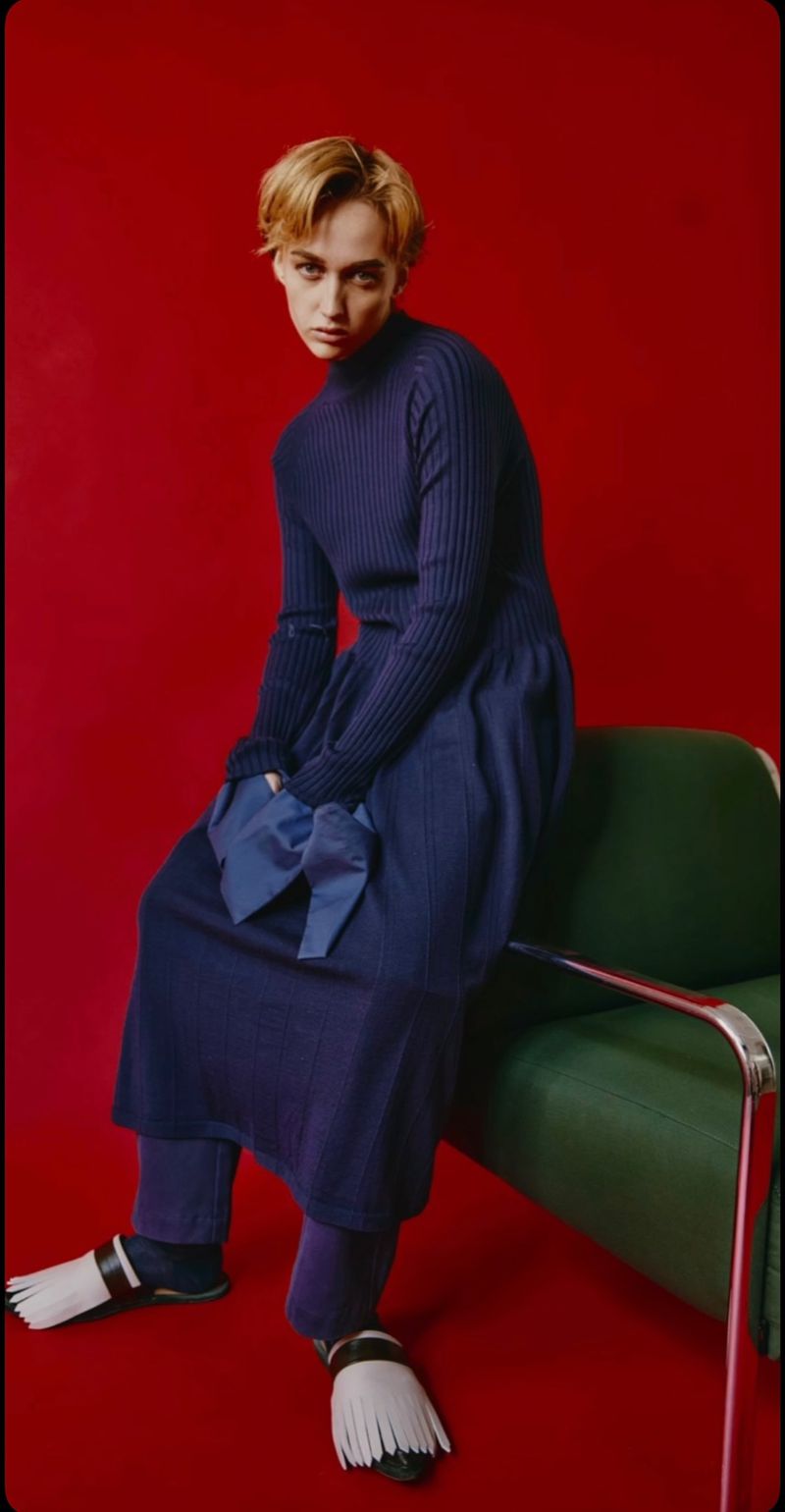 Fashion portrait of a non-binary person sitting on a green chair against a red background wearing a navy blue dress with fringe shoes.