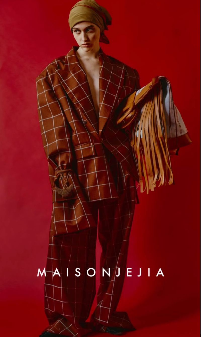 A fashion image shows an adult female model wearing a brown and white checkered suit with a headscarf, posed against a solid red backdrop.