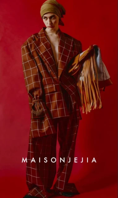 A fashion image shows an adult female model wearing a brown and white checkered suit with a headscarf, posed against a solid red backdrop.