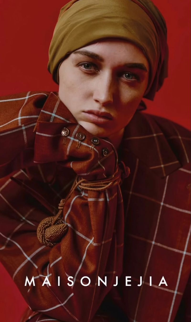 Close-up of a woman wearing a brown and white plaid jacket and tan headscarf against a red background, fashion campaign.