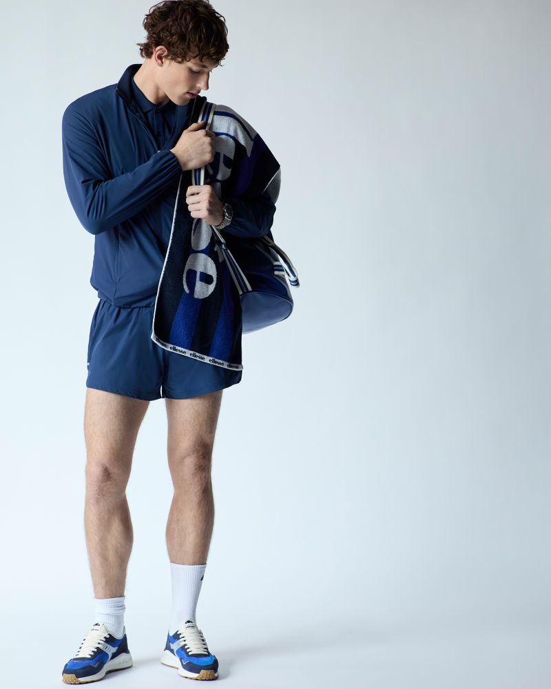 Athletic man in blue tracksuit and white socks posing with a branded towel, wearing running shoes indoors, studio lighting.
