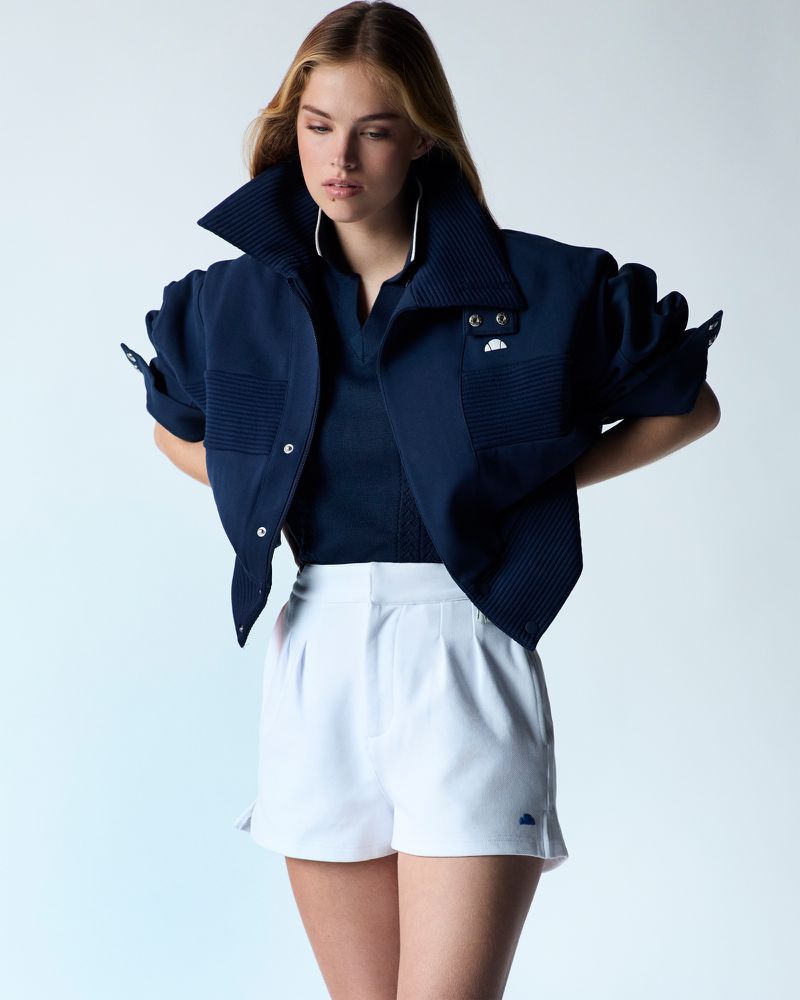 A young woman in a blue jacket and white shorts poses in a studio with soft lighting, showcasing a modern and stylish fashion look.