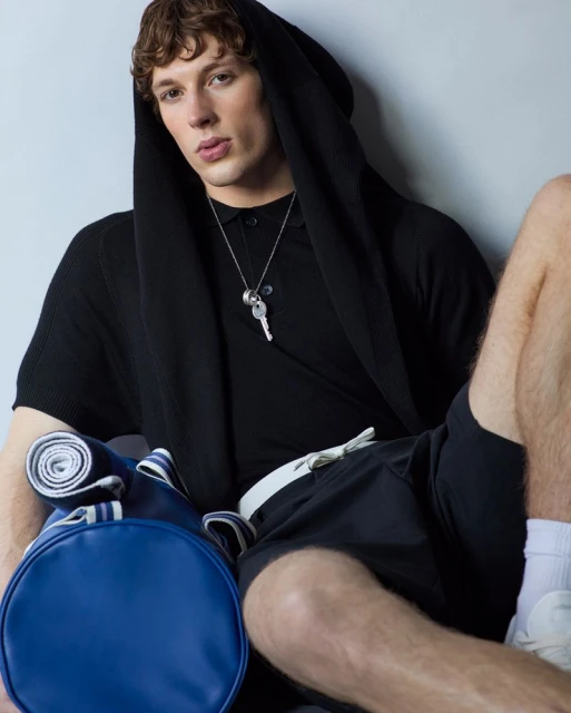 Man in black hooded shirt and shorts sitting with a blue duffel bag, wearing a key necklace, indoors against a neutral wall.