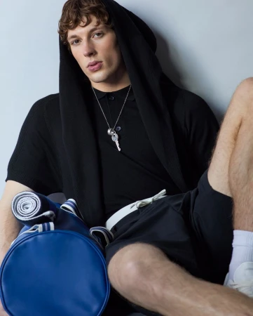 Man in black hooded shirt and shorts sitting with a blue duffel bag, wearing a key necklace, indoors against a neutral wall.