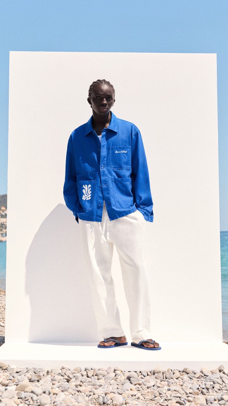 A dark-skinned male fashion model poses outdoors on a rocky beach in a blue jacket and white pants in front of a white screen.