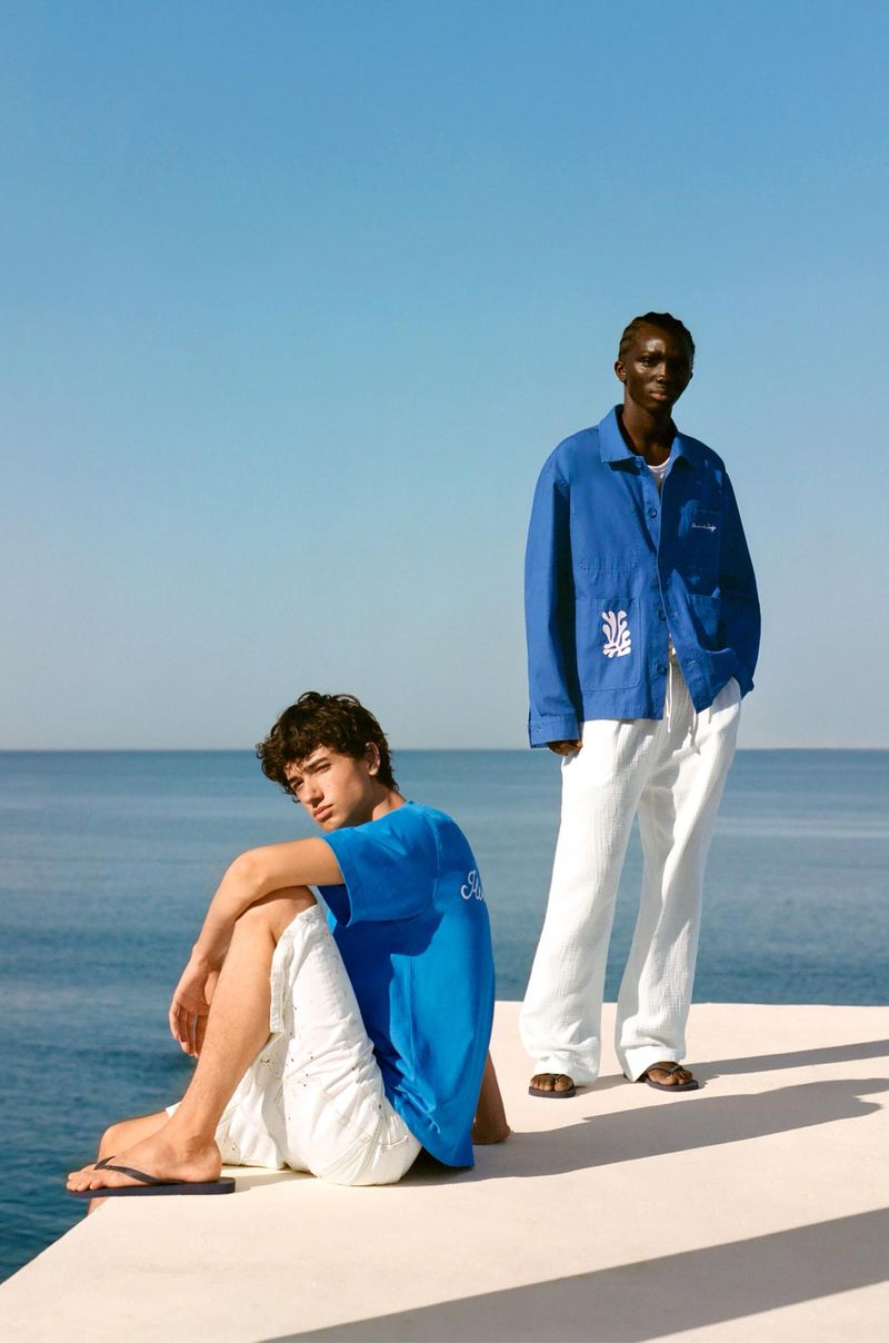 Two men in a bright, outdoor fashion shot. One seated, wearing a blue tee and white shorts. The other stands, wearing a blue jacket and white pants.