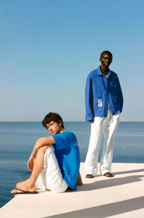 Two men in a bright, outdoor fashion shot. One seated, wearing a blue tee and white shorts. The other stands, wearing a blue jacket and white pants.