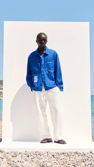 Model Abdoulaye Fall in a bright blue overshirt and loose white trousers standing before a white backdrop on a rocky beach.